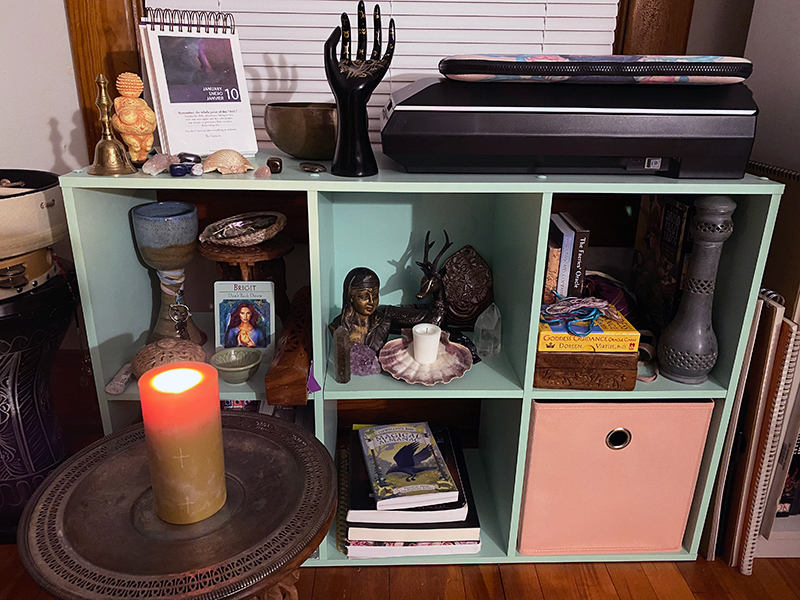 A low bookshelf with cube shaped openings, being used as a Wiccan altar, with various decorations, statues, books, and stones.