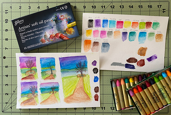 A photo of two sheets of watercolor paper with thumbnail planning sketches on one and color swatches on the other. There are oil pastel boxes above and below the papers.