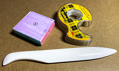 A small three-dimensional paper box next to double-sided tape and a bone folder
