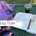 Photo of a green, metal picnic table. Ontop of the table is a an open sketchbook with drawings of trees, an plastic cup of iced coffee, a purple backpack, and a pencil case with art pencils and erasers.