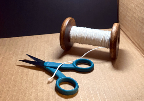 Photo of an antique wooden spool and a small pair of scissors with teal plastic handles, inside a cardboard box and lit from above by a bright light.